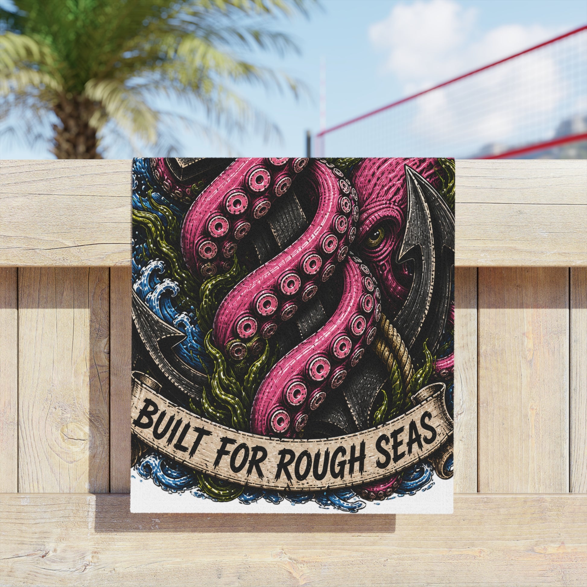 Nautical beach towel with kraken tentacles and anchor design, hanging on a timber fence under a palm tree.